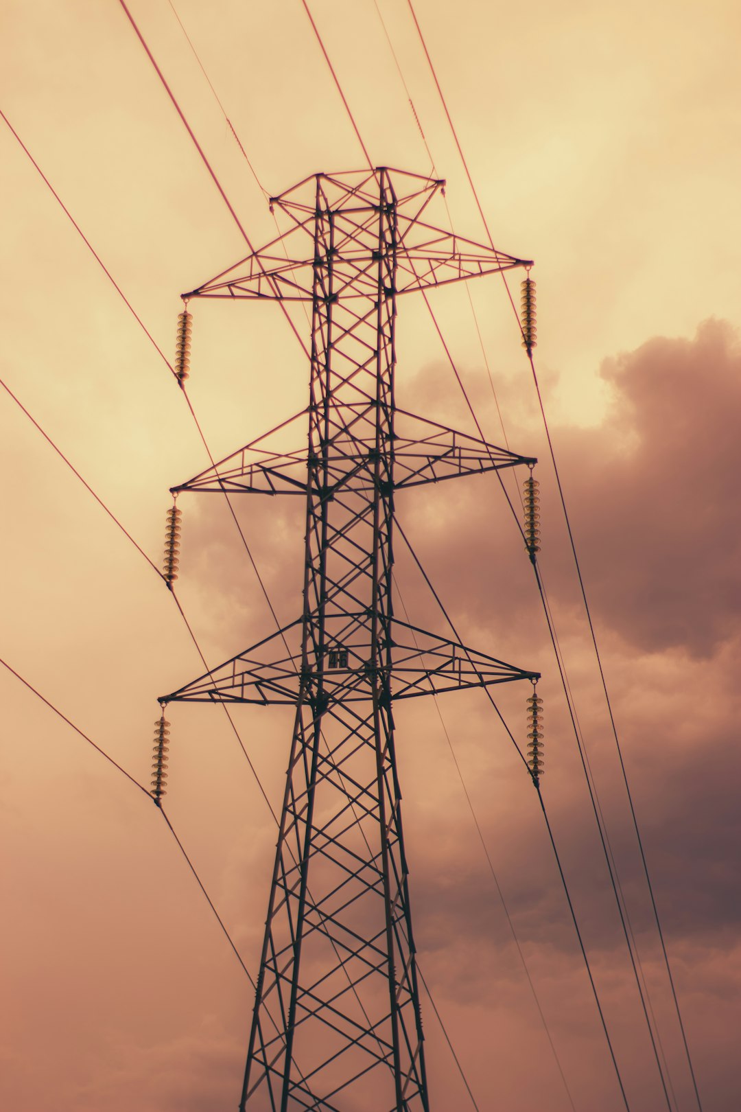 Lattice high-voltage power transmission tower with multiple lines against a warm sunset sky underneath cloudy horizon