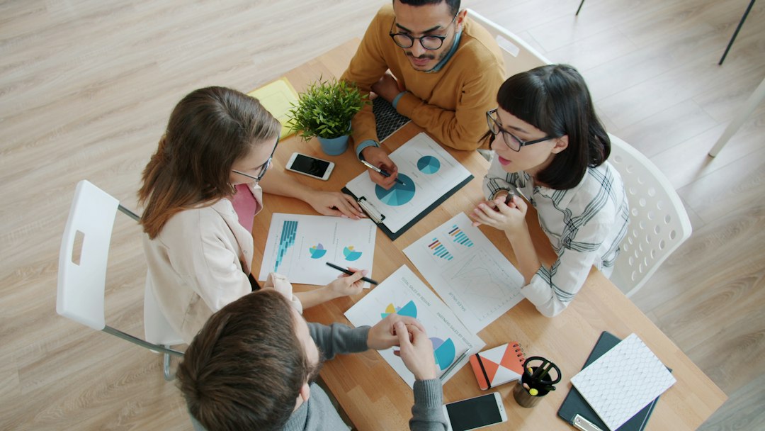Four coworkers sit around a wooden table, reviewing charts and graphs on printed reports together.",