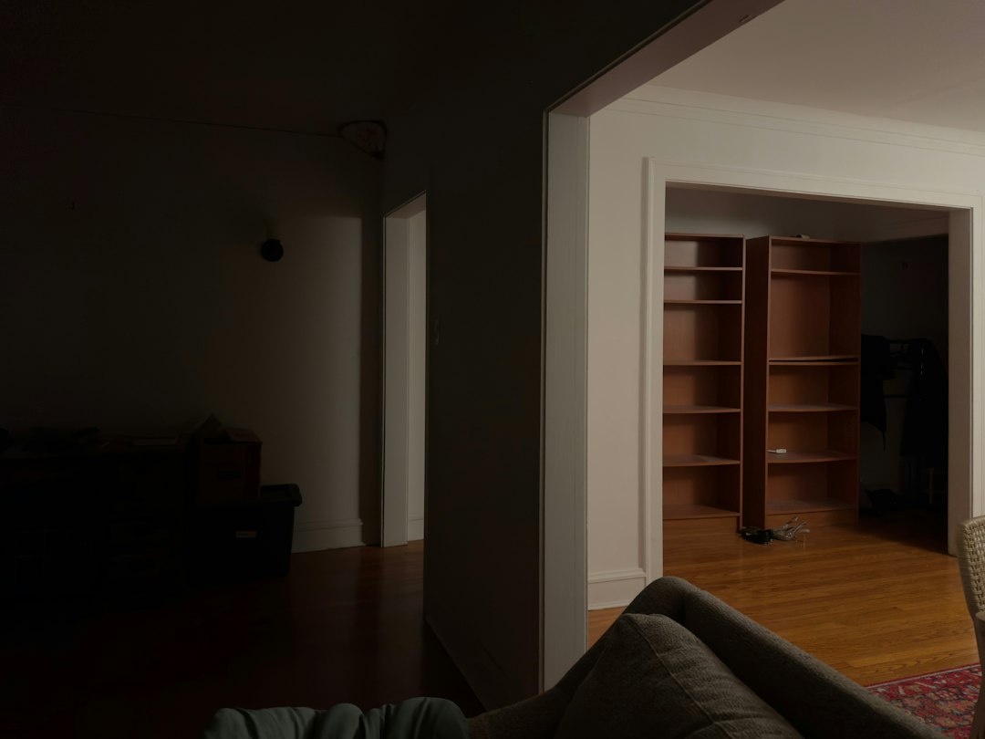 Dim living room with a foreground sofa; an open doorway reveals a room with tall empty wooden bookshelves and a hardwood floor.
