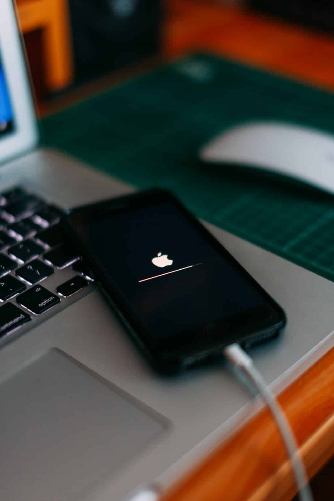 Black iPhone plugged in to a silver laptop, showing an Apple logo with a progress bar on screen as it charges/updates.