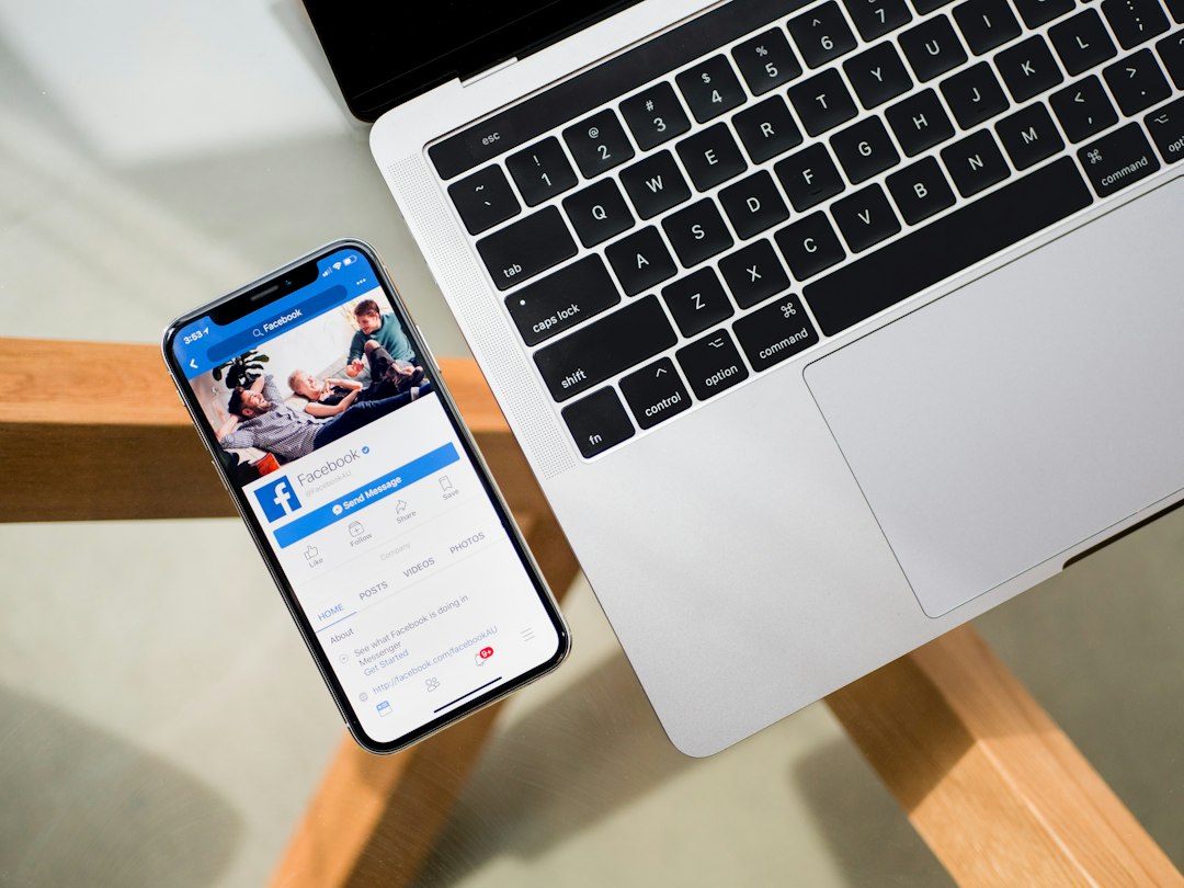 Laptop keyboard beside a smartphone displaying the Facebook app open on a wooden table surface