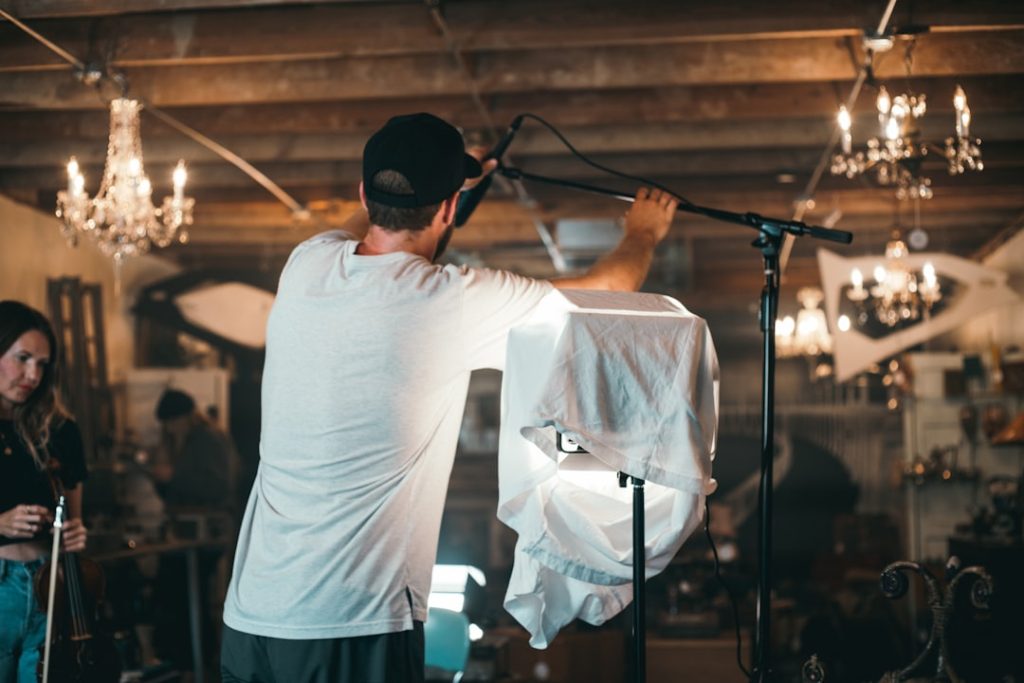 A man in a white t-shirt adjusts a microphone on a stand in a cozy, chandelier-lit studio while a woman with a violin watches nearby.