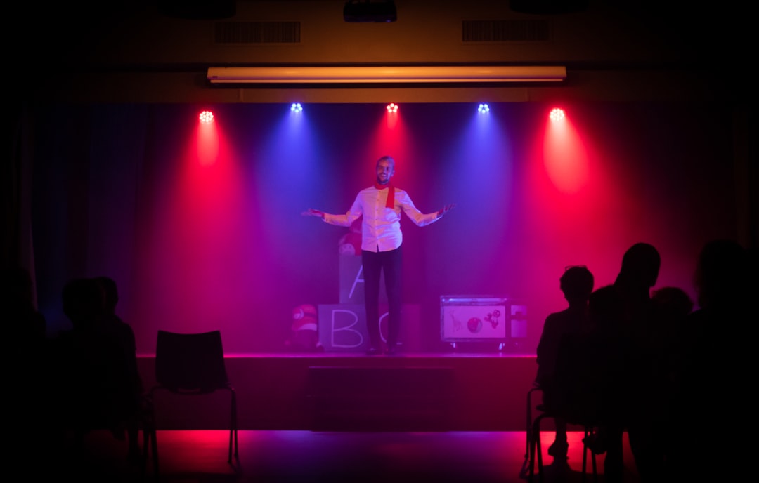 Performer on a dimly lit stage with red and blue spotlights, arms outstretched, audience silhouettes in the foreground.