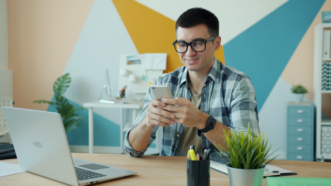 Man in plaid shirt and glasses smiles while using a smartphone at a tidy desk with a silver laptop nearby in a colorful office.