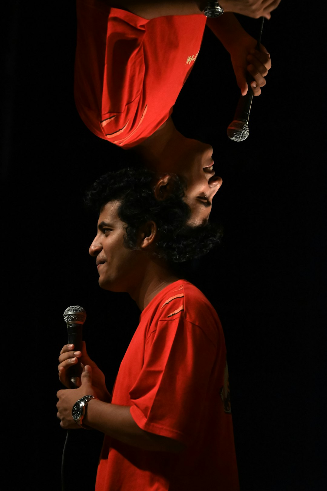 Two mirrored images of a man in a red shirt holding a microphone on a dark stage, speaking to an audience.