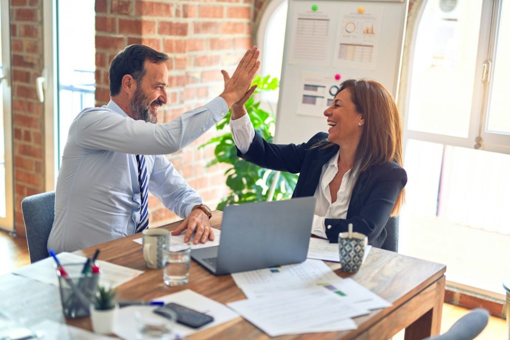 Two professionals celebrate with a high-five across a wooden desk, laptop open and papers scattered nearby in a bright office.