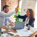 Two professionals celebrate with a high-five across a wooden desk, laptop open and papers scattered nearby in a bright office.