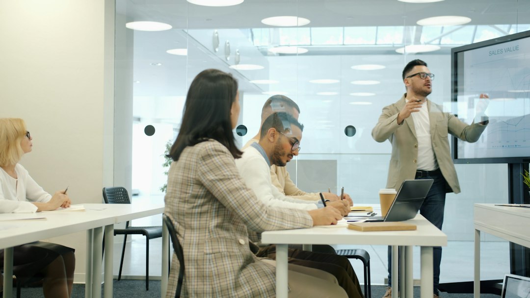 A presenter in a beige blazer explains a chart on a large screen to a conference room group, with colleagues taking notes at white tables.