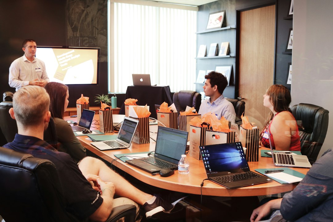 Presenter speaks to a group in a conference room as attendees work on laptops and orange gift bags line the table.