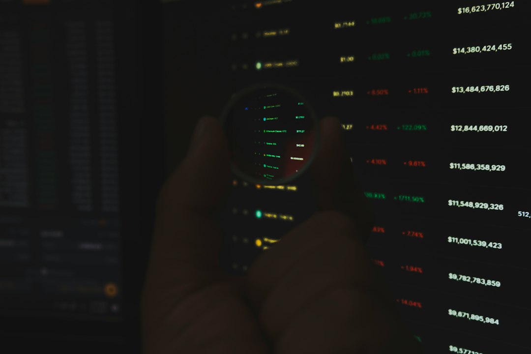 Hands holding a magnifying glass over a dark screen showing green and red financial numbers and data rows.