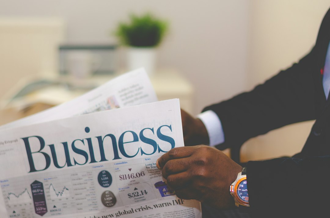 Person in a suit reads a newspaper with a large 'Business' headline in an office setting, a wristwatch visible on the arm.