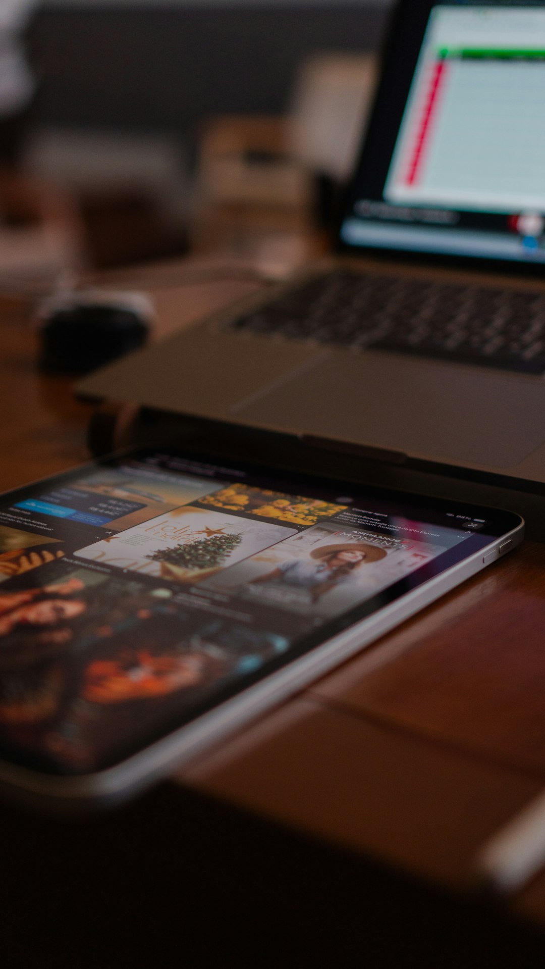 Tablet on a wooden desk shows a magazine app with colorful covers; a laptop sits blurred in the background.