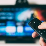 Hand holding a black remote control pointed at a blurred television screen in the background.
