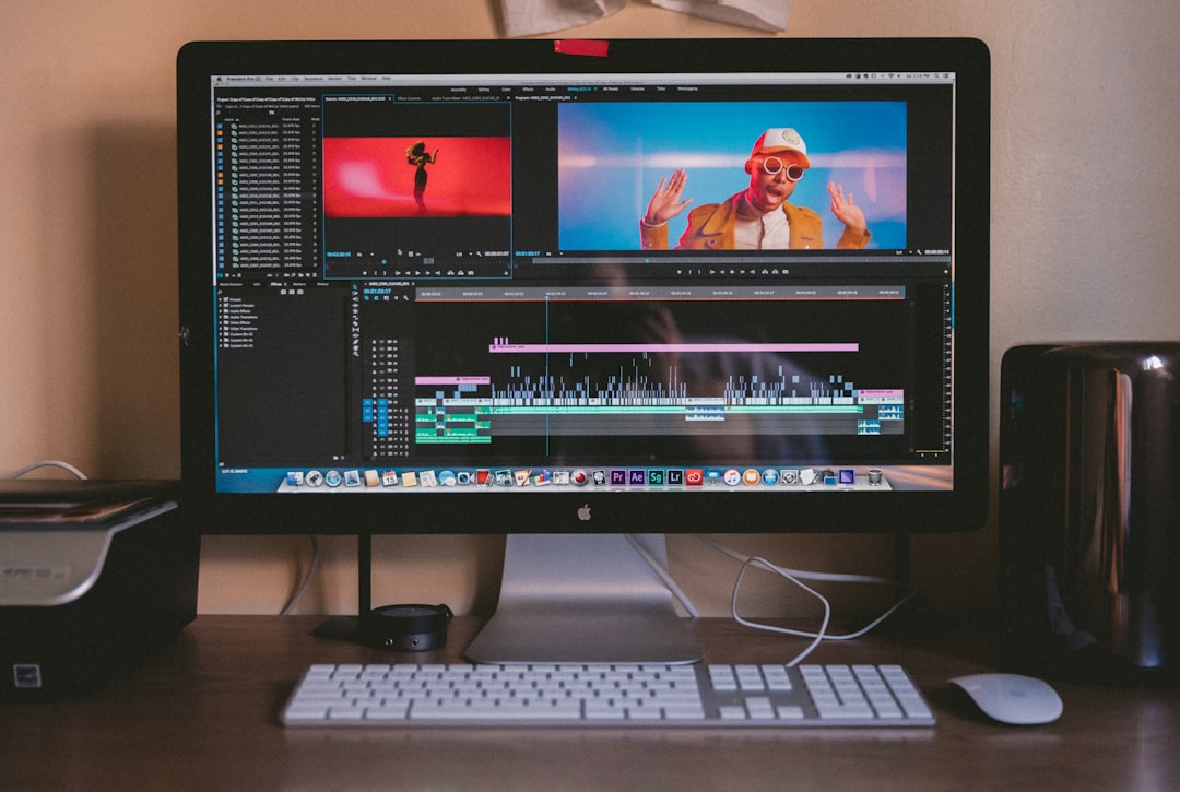 Desktop monitor showing video editing software with a timeline, two preview panels, and colorful clips on the timeline; a keyboard and mouse on a wooden desk.