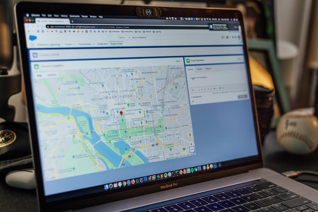 Laptop screen showing an online map with a red location pin and surrounding streets, on a desk beside a baseball and gadgets