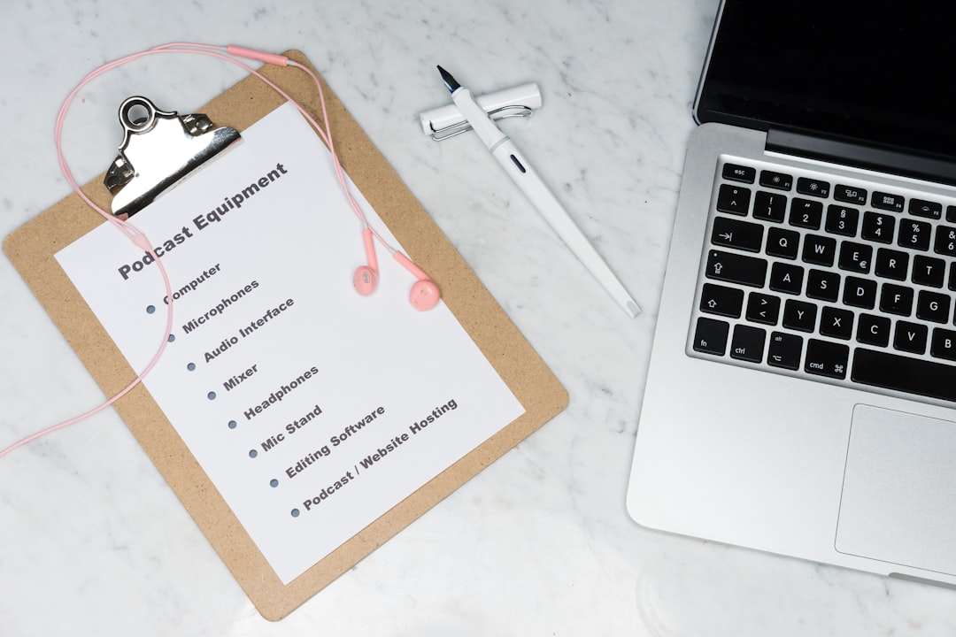 Clipboard labeled 'Podcast Equipment' with a gear checklist and pink earbuds on a marble desk beside a laptop inside view of a workspace.