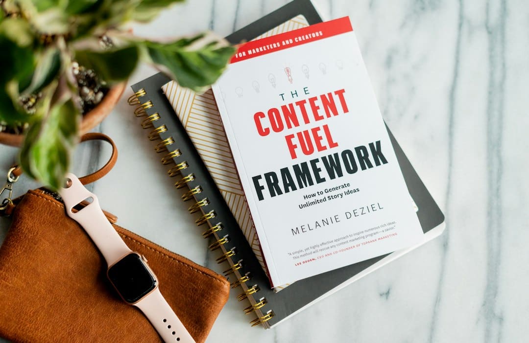 Book titled The Content Fuel Framework on a desk with a spiral notebook, plant, and pink smartwatch.