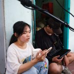 Young woman sits outside a doorway, holding a cup and looking at her phone while a man next to her uses a tablet.