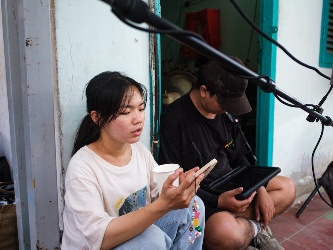 Young woman sits outside a doorway, holding a cup and looking at her phone while a man next to her uses a tablet.