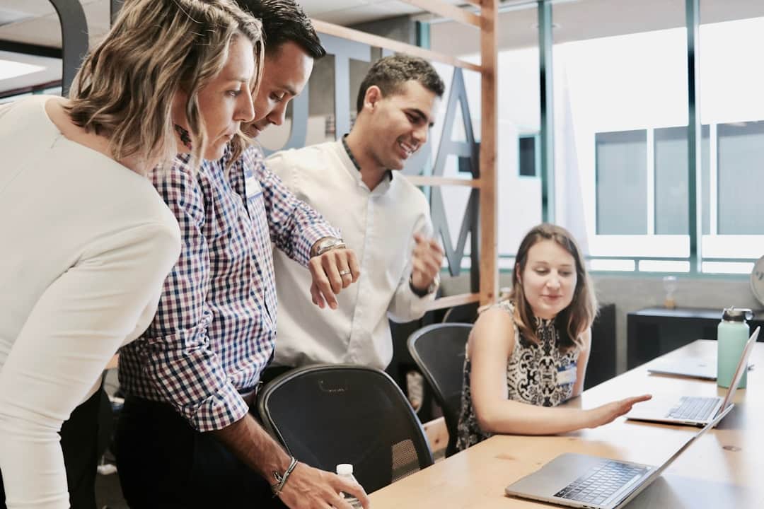 A group of colleagues gathered around a laptop, discussing a project in a modern office setting.