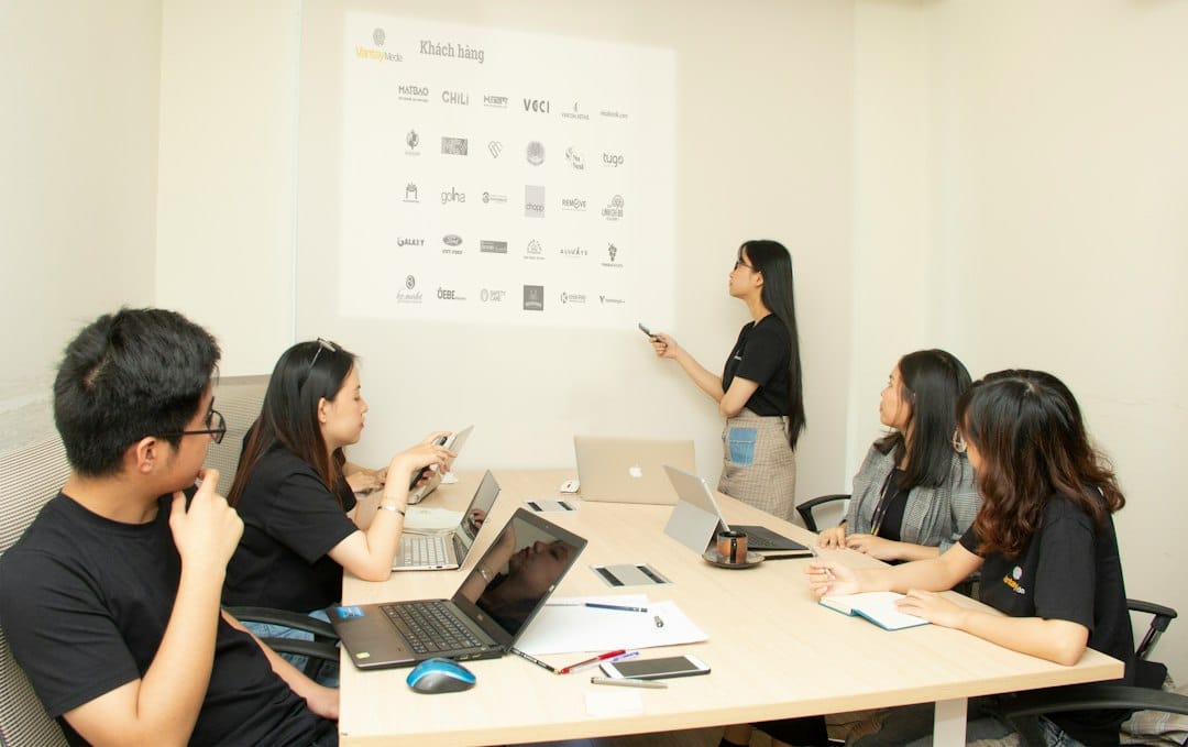 Presenter points to a wall screen displaying logos while a team of five sits around a conference table with laptops and notes nearby.