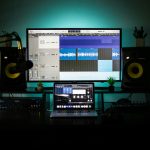 Home recording studio setup with a large monitor showing a DAW, a laptop, studio monitors, and teal backlight behind.