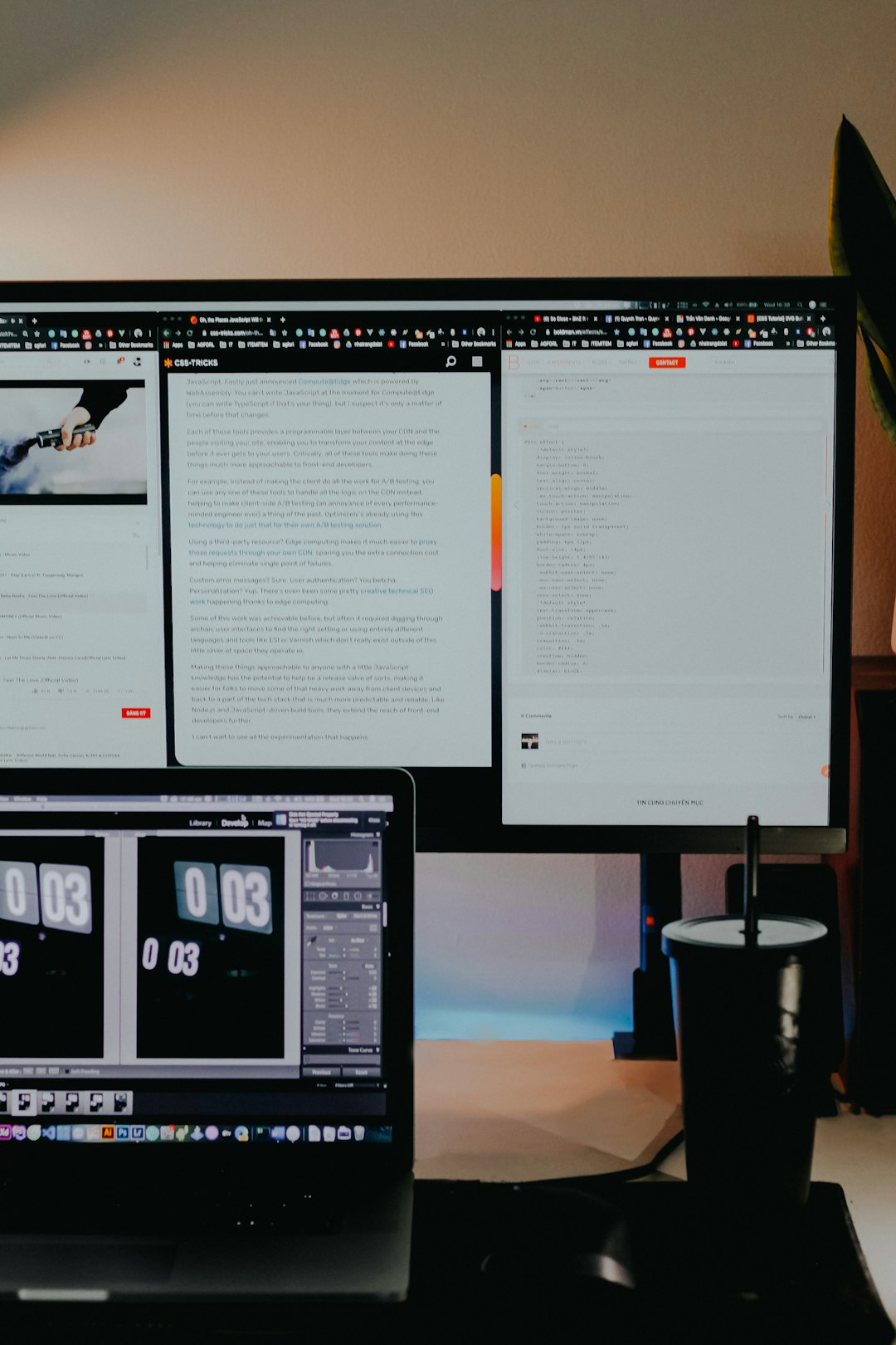 Large monitor on a desk showing multiple open windows, with a laptop below and a black cup on the right side.