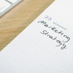 Calendar page showing date 23, with handwritten 'Marketing Strategy' on a dotted planner beside a wooden desk and a white keyboard edge in the corner.