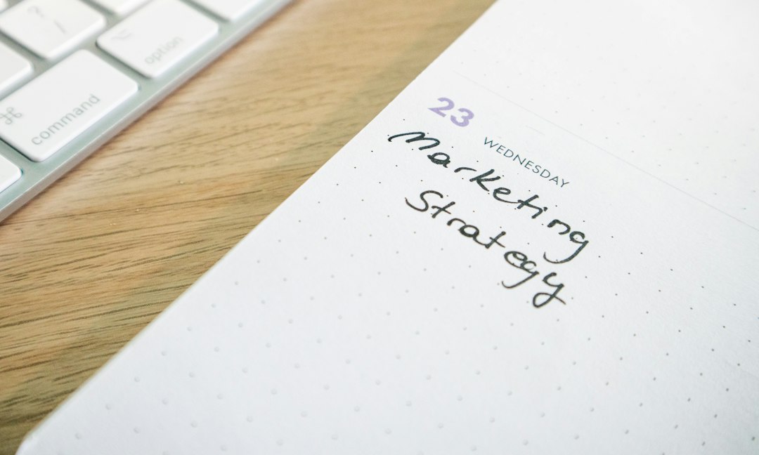 Calendar page showing date 23, with handwritten 'Marketing Strategy' on a dotted planner beside a wooden desk and a white keyboard edge in the corner.