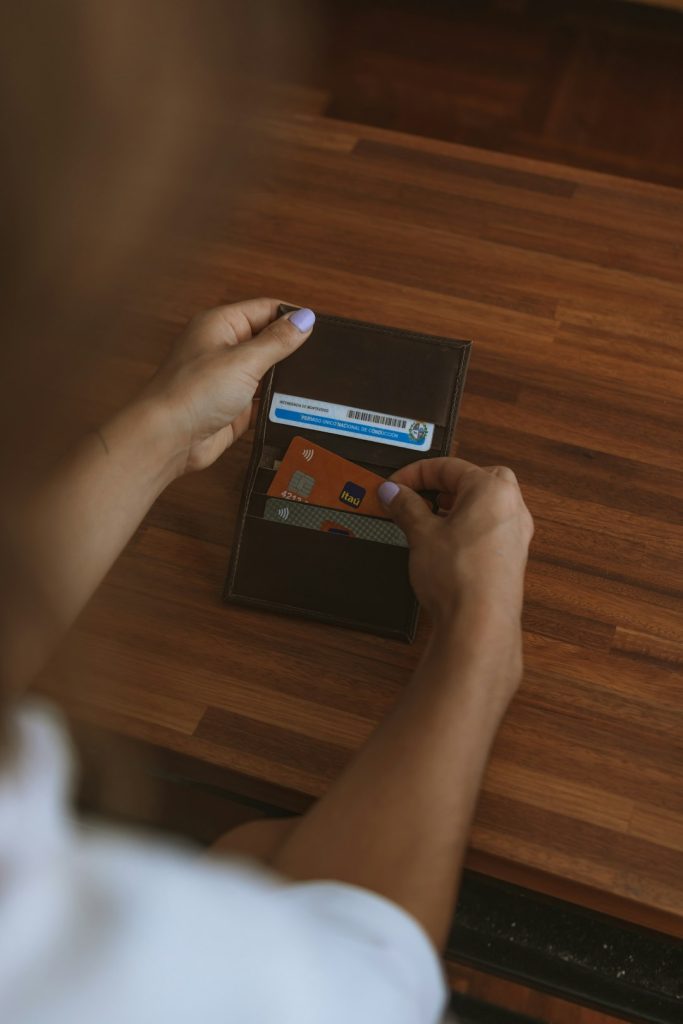 Person’s hands hold a brown leather wallet on a wooden table, exposing several cards in the card slots.