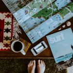 Top-down view of a travel planning setup: open map on a wooden table with a cup of coffee, shells, a light blue surf magazine, and a smartphone showing a photo; feet visible at bottom edge.