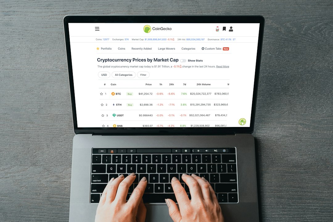 Laptop on a wooden desk displaying a CoinGecko crypto prices dashboard with BTC, ETH, USDT rows visible.