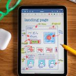 Digital wireframe sketch of a landing page on a tablet screen, with annotated notes, a pencil nearby, and a pair of green glasses on a wooden desk.