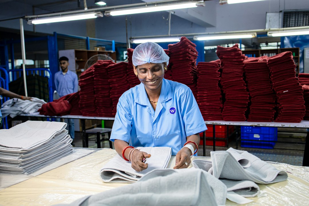 Factory worker in blue uniform folds light gray textiles at a table with stacks of red folded fabrics in the background.