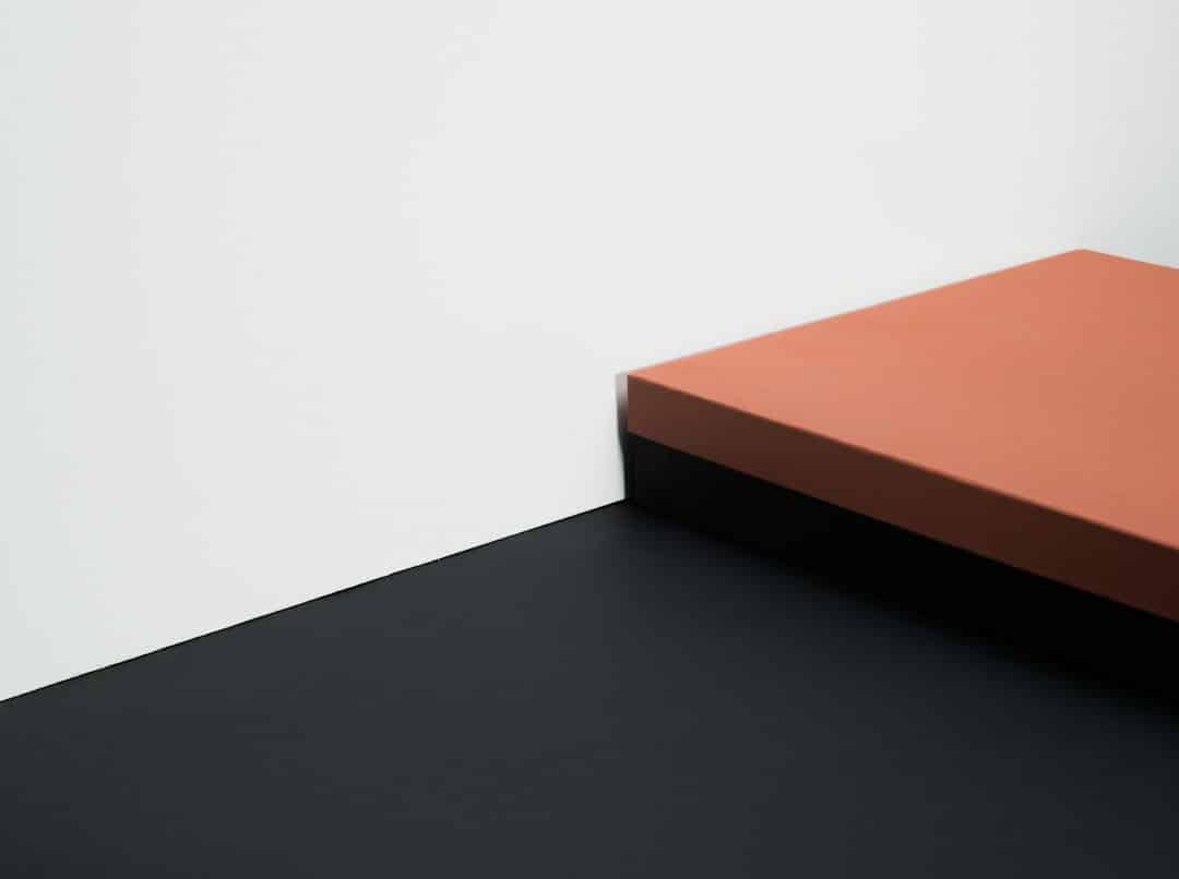 Orange rectangular platform jutting over a dark floor at a wall corner, photographed in a minimalist style