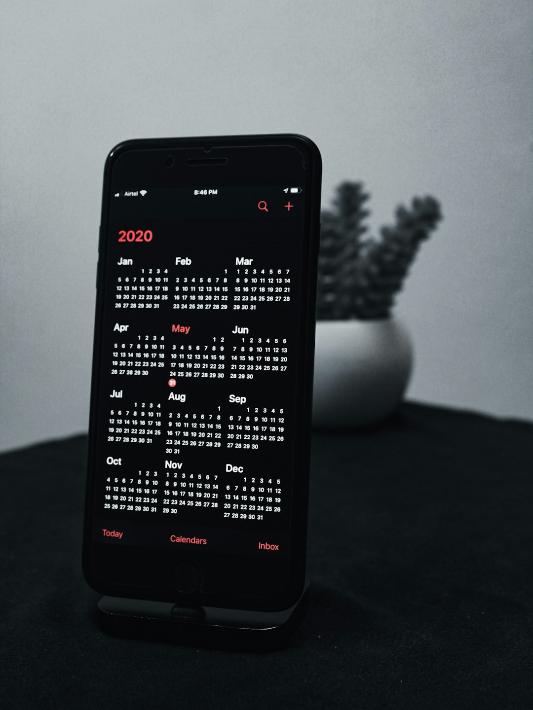 Smartphone showing a 2020 calendar app with monthly grids on a dark screen, resting on a dark surface with a blurred plant in the background.
