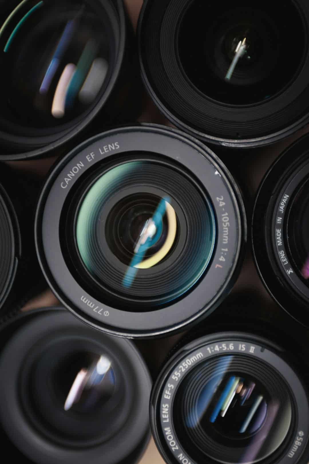 Close-up of multiple camera lenses stacked together, with the foreground lens showing 'CANON EF LENS 24-105mm' and colorful reflections on the glass surface