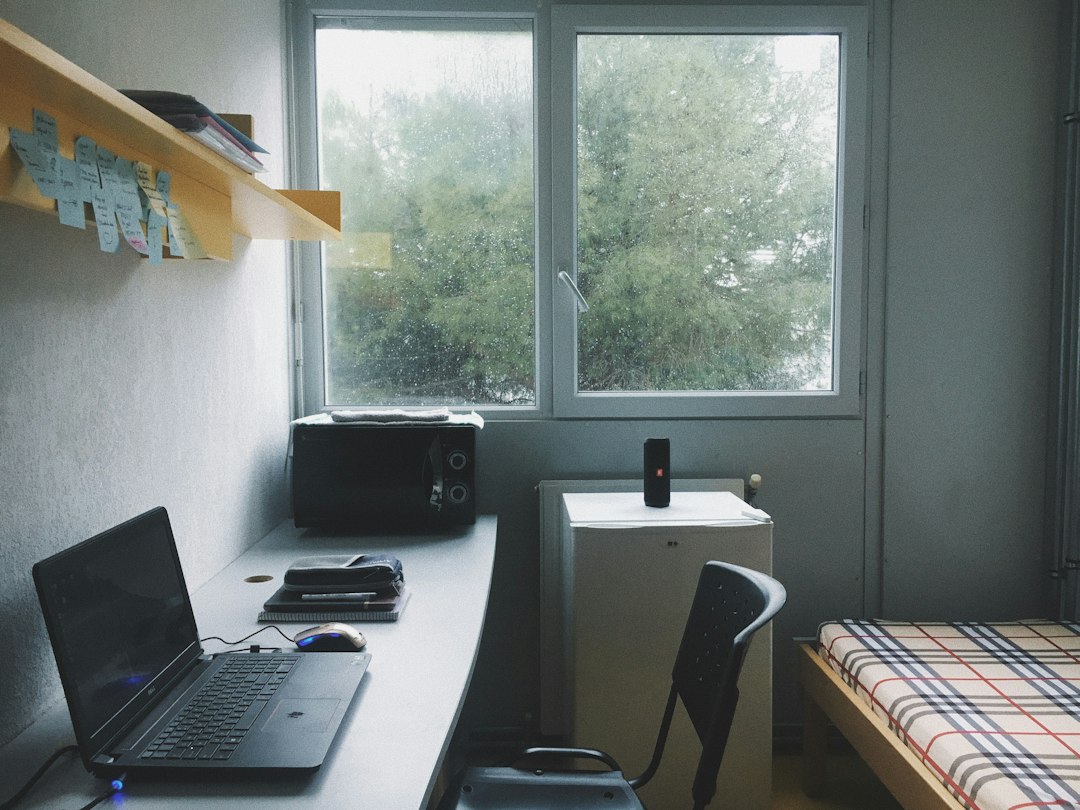 Dorm room workspace with a laptop on a white desk, mouse, notebooks, and a small microwave against a windowed wall; a mini fridge sits near a black office chair, and a bed with a plaid blanket is visible to the right.
