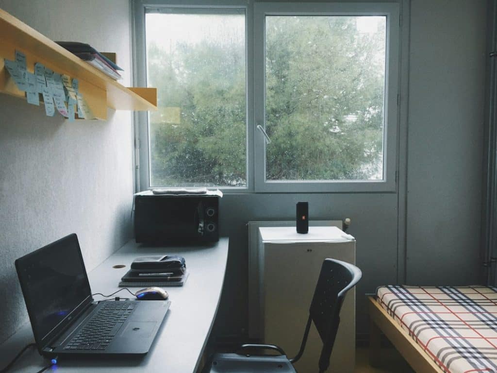 Dorm workspace: laptop on a long desk with a mouse, notebooks, and a microwave; a small fridge sits under a window, and a plaid-bed is on the right.