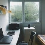 Dorm workspace: laptop on a long desk with a mouse, notebooks, and a microwave; a small fridge sits under a window, and a plaid-bed is on the right.