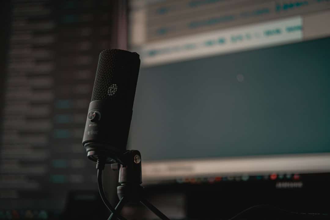 Close-up of a black condenser microphone mounted on a stand, with a computer monitor blurred in the background.