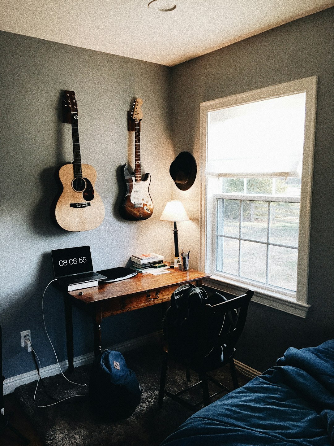 Corner bedroom workspace with two guitars hanging on the wall above a wooden desk, a laptop showing the time 08:08:55, a lamp, books and stationery on the desk, and a chair facing a window letting in daylight.