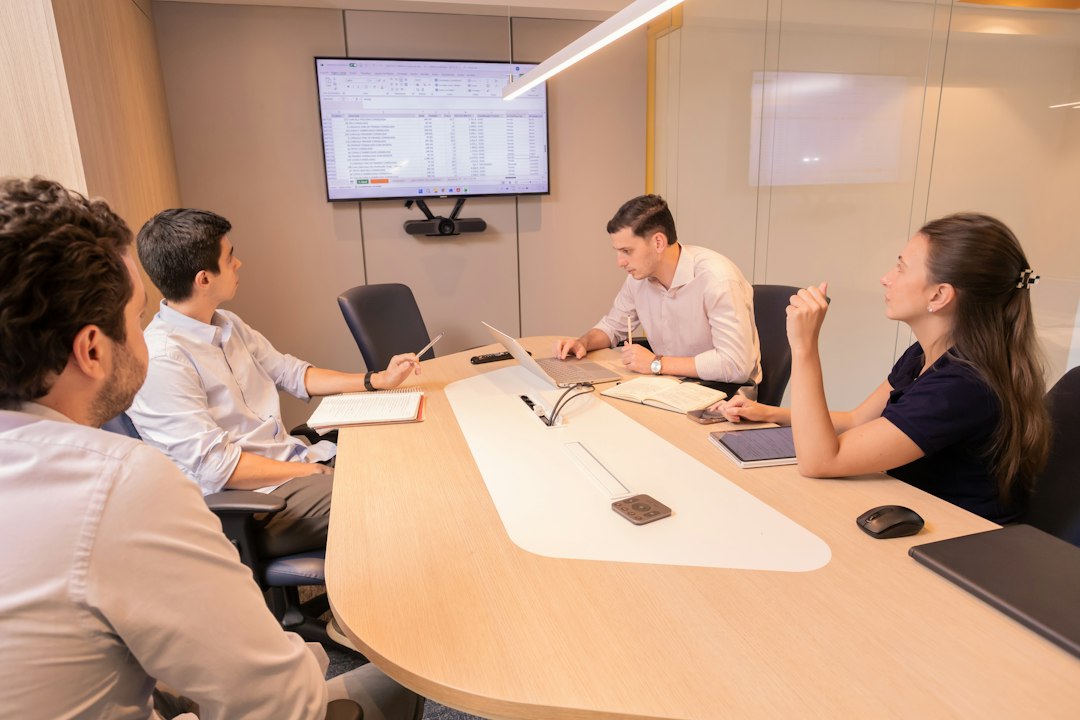 Four professionals in a modern conference room discuss around a curved wooden table, with a large screen displaying spreadsheet data behind them