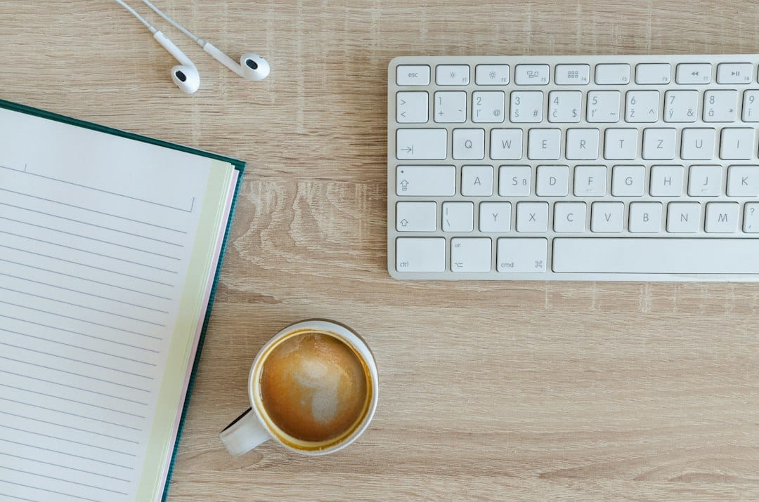 Top-down view of a workspace: a white keyboard on a light wood desk, with an open notebook on the left and a cup of coffee in the foreground, plus white earbuds at the top left.