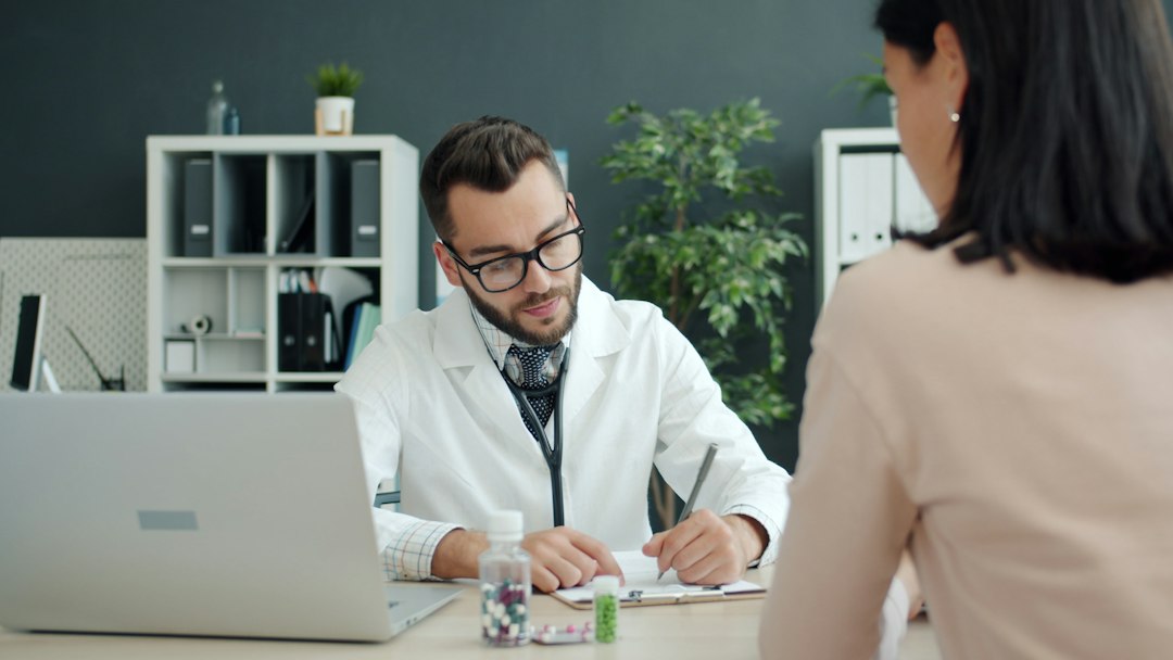 Doctor with glasses and a stethoscope reviews a form during a consultation with a patient in a modern clinic office.