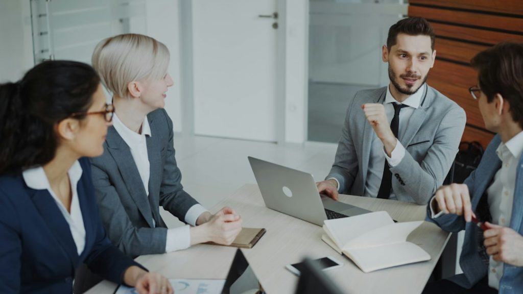 Four professionals in a business meeting around a table office meeting silent agreement workplace communication - AI Bud Four professionals in business attire sit around a conference table in a modern office as a man in a gray suit gestures while others listen.