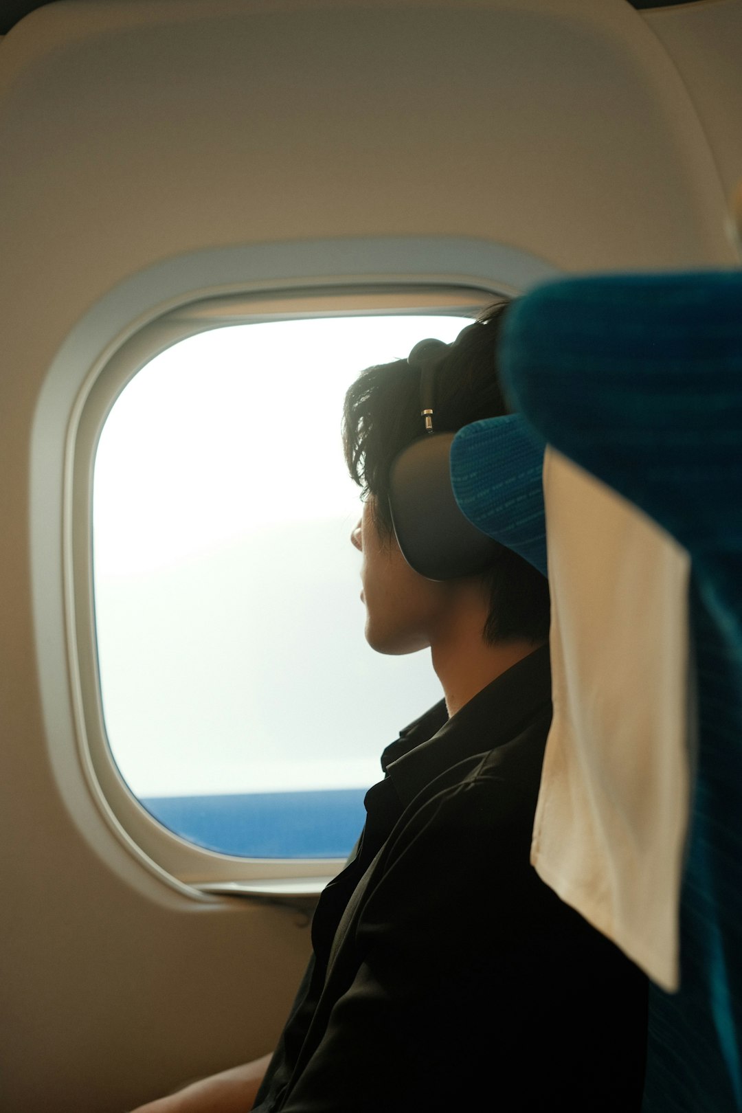 Traveler wearing over-ear headphones gazing out airplane window at the ocean beneath the winged horizon/sea view.