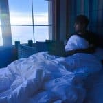 Young man sits up in a white duvet, using a laptop by a large window with a mountain view outside.