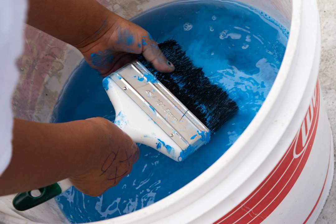 Hands hold a paintbrush dripping blue paint over a large white bucket of bright blue paint, ready to paint.