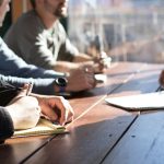People sit around a wooden table, writing in notebooks during a meeting or discussion.